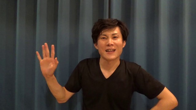 青いカーテン背景で手を挙げて話す本人（動画「トラブルを人のせいにしたら赤信号です」より）
