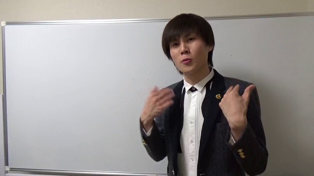 ホワイトボード前でスーツにネクタイピン姿の本人が両手を広げて話す（動画「お金持ちに会うメリット【パチスロ】【スロット】」より）