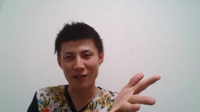 無地の壁を背景に花柄シャツ姿の本人がピースサインをして話す（動画「最近のパチスロで稼げる人になるには？【パチスロ】…」より）