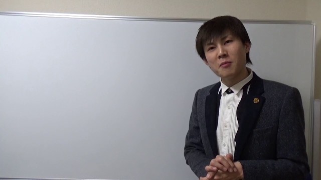 スーツにネクタイ姿でホワイトボード前に立つ本人（動画「借金返済は頑張る理由にならない【パチスロ】…」より）