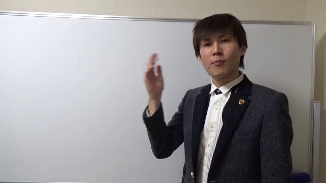 グレースーツでホワイトボード前に立ち片手を挙げる本人（動画「悩めるスロッターへ【パチスロ】【スロット】」より）