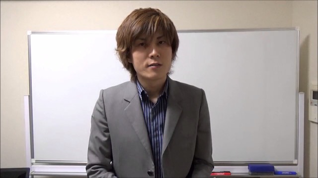 縞シャツにグレースーツでホワイトボード前に立つ本人（動画「自己流は良くない」より）