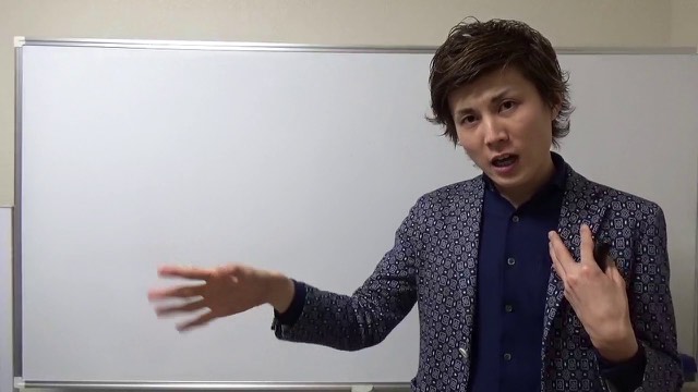 柄物ジャケットでホワイトボード前に立ち片手を伸ばす本人（動画「金持ちになった人がしてた事【パチスロ】【スロット】」より）