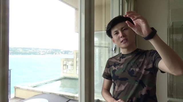 海の見える窓際で迷彩Tシャツを着て話す本人（動画「【答え】パチスロや投資で勝てない理由」より）