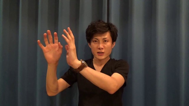 青いカーテンを背景に両手を挙げる黒いVネック姿の本人(動画「チャンスの神様は前髪しかない これは正しいです」より)