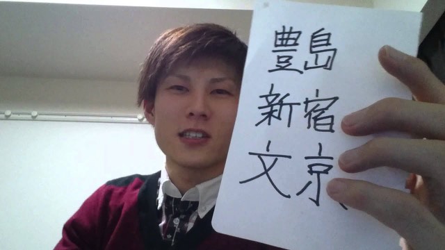 「豊島　新宿　文京」と書いた紙を顔の横に掲げる本人（動画「12月稼働地域抽選」より）